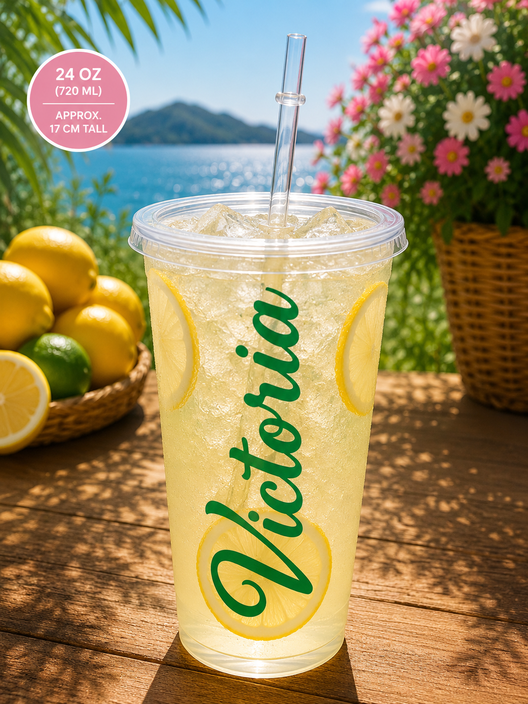 A clear 24 oz plastic cup with a clear straw, filled with a light yellow iced drink and decorated with lemon slice graphics, featuring the name 'Victoria' in green script. The cup is set on a wooden table with a sunny outdoor background showing flowers, lemons, and water.
