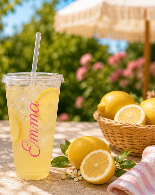 Yellow lemonade in a clear cup with a straw, placed on a wooden surface with lemons and a basket in the background.