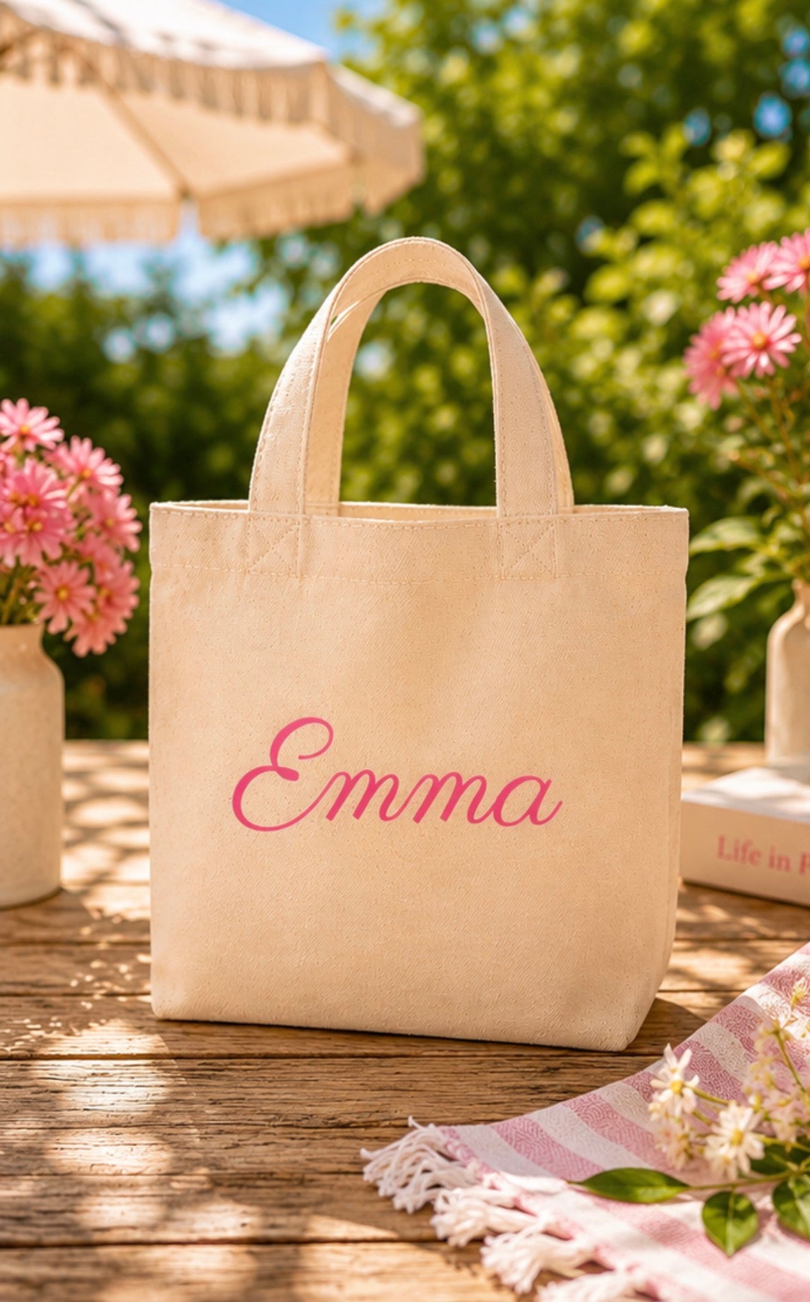 Beige tote bag with 'Emma' embroidered on it, placed outdoors on a wooden surface with flowers and an umbrella in the background.