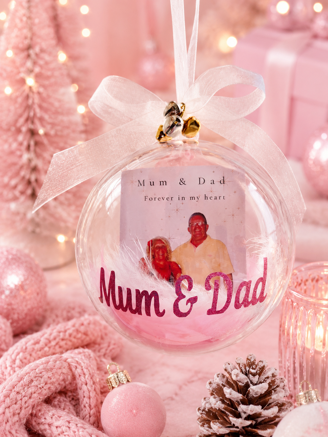 A round clear Christmas ornament with a white ribbon bow and small gold bells, featuring a printed photo of a couple and the words 'Mum & Dad Forever in my heart' and 'Mum & Dad' in pink script. The ornament is photographed in a soft pink holiday setting with lights, wrapped gifts, a knit scarf, and a pinecone.