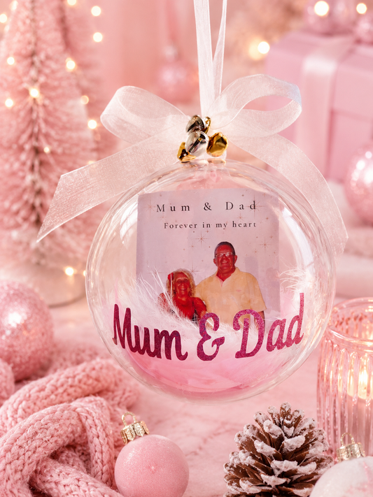 A round clear Christmas ornament with a white ribbon bow and small gold bells, featuring a printed photo of a couple and the words 'Mum & Dad Forever in my heart' and 'Mum & Dad' in pink script. The ornament is photographed in a soft pink holiday setting with lights, wrapped gifts, a knit scarf, and a pinecone.