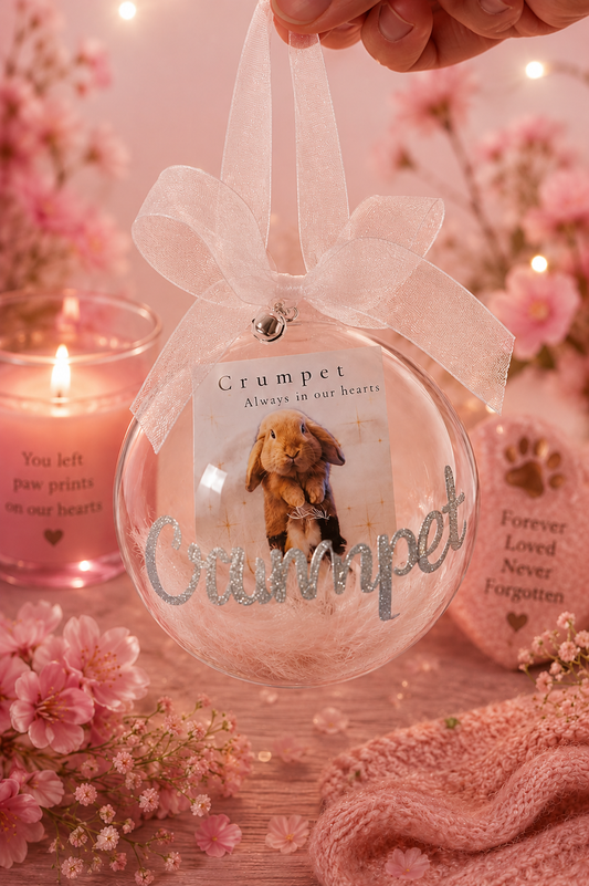 A clear round hanging ornament with a white ribbon bow, featuring a printed bunny photo, the name 'Crumpet' in silver glitter, and a small card that reads 'Crumpet Always in our hearts.' A hand holds the ornament, surrounded by pink flowers, a lit candle, and a knitted pink scarf in a soft, warm setting.