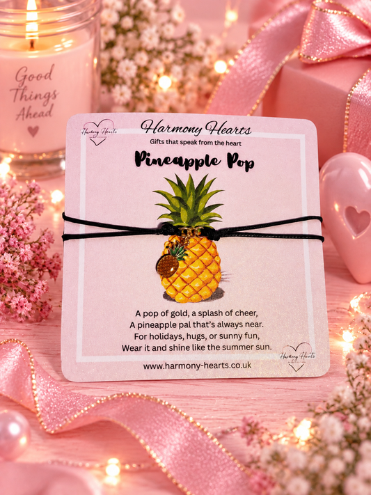 A square card reading 'Harmony Hearts Pineapple Pop' with a pineapple charm on a black cord bracelet, placed on a pink surface with flowers, a candle, and pink ribbon around it. The card features a pineapple illustration and a short poem.