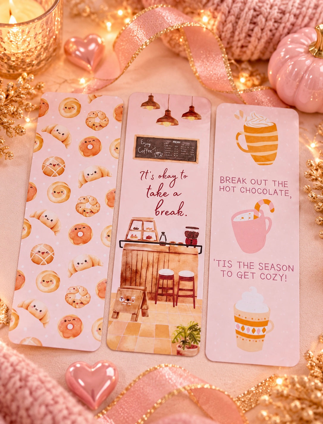 Three pink decorative bookmarks styled on a cozy tabletop with warm fairy lights, ribbons, and hot chocolate-themed props. The bookmarks feature illustrated donuts, a cozy cafe interior with the quote 'It's okay to take a break.