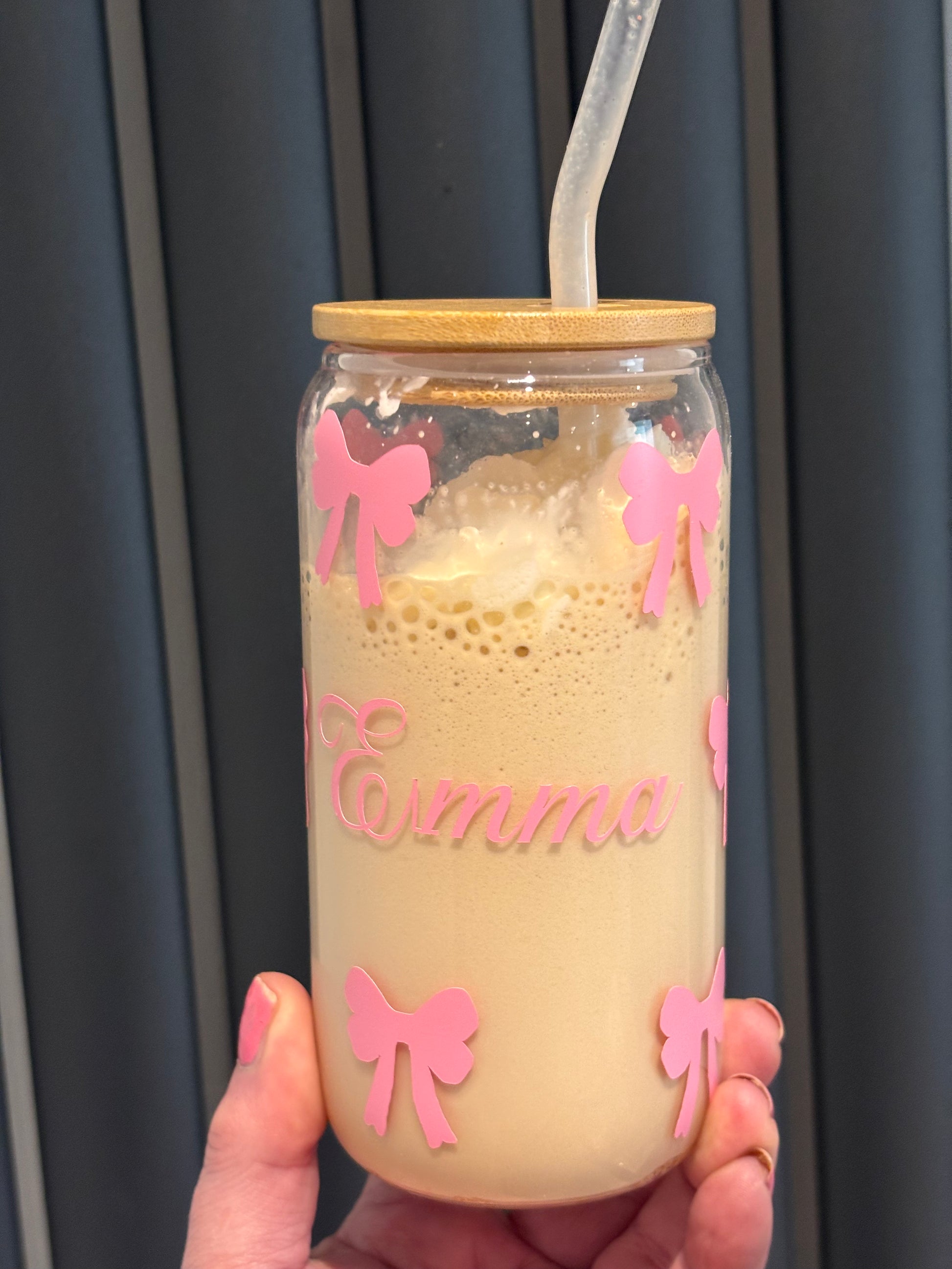 Glass tumbler with wooden lid and pink bows, containing a creamy beverage, held by a hand against a dark curtain background.