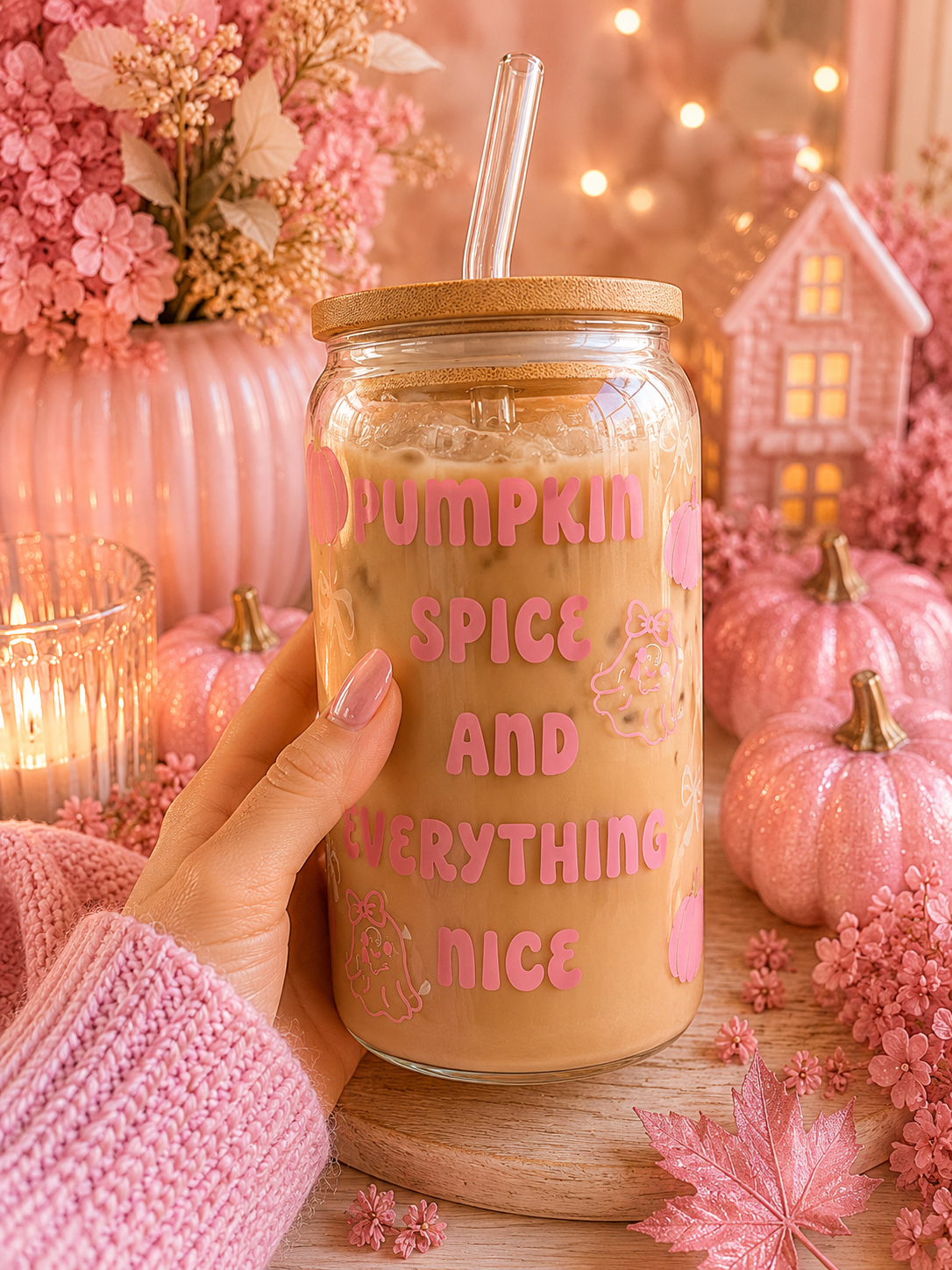 A hand in a pink knit sweater holds a clear glass jar with a bamboo lid and clear straw, filled with an iced coffee drink. The jar has pink lettering that reads 'PUMPKIN SPICE AND EVERYTHING NICE' with small floral illustrations.