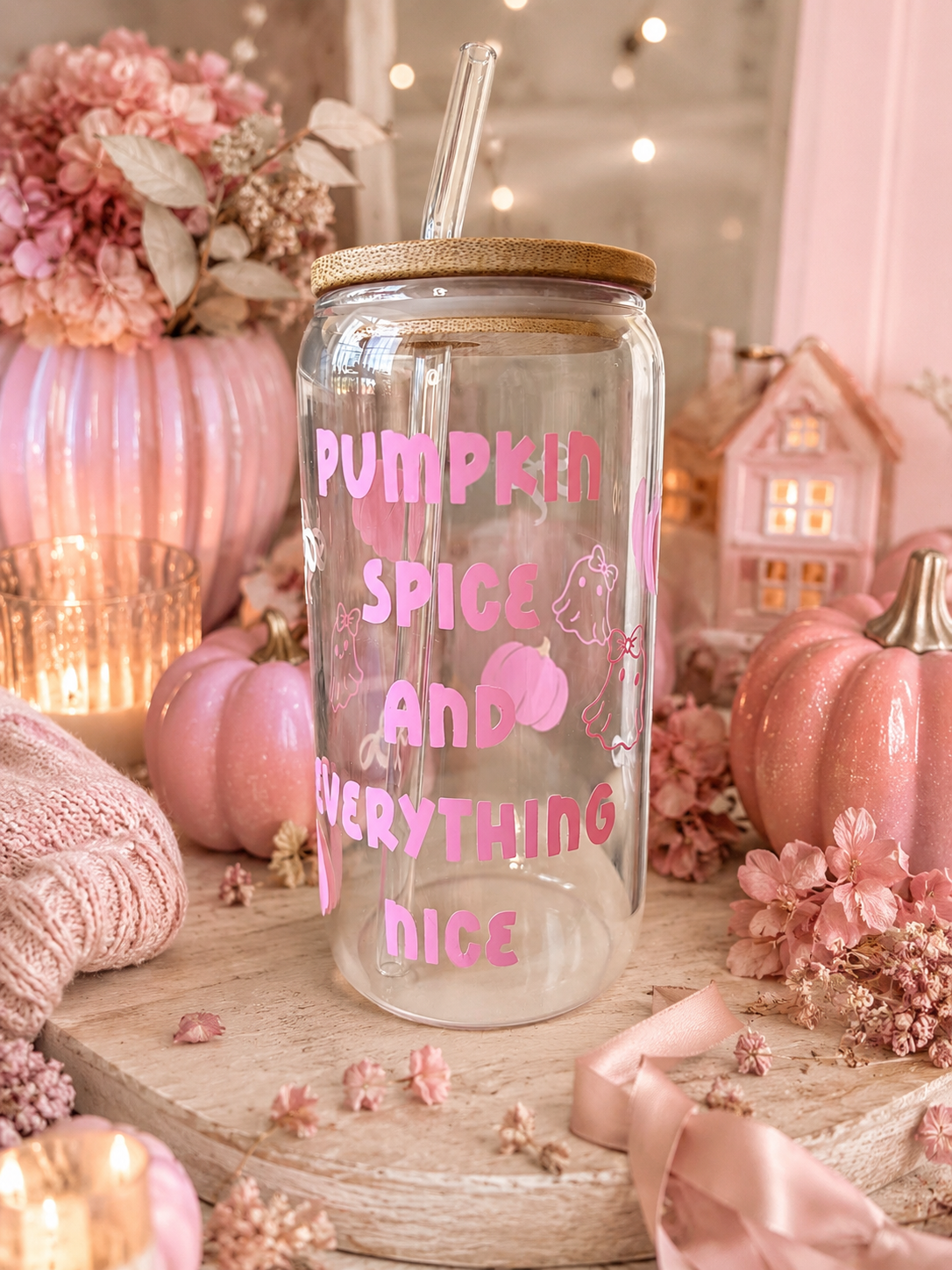 A clear glass mason jar tumbler with a bamboo lid and clear straw, decorated with pink text reading 'PUMPKIN SPICE AND EVERYTHING NICE' and a pink pumpkin graphic. The jar is styled in a cozy autumn setting on a wooden board with pink pumpkins, dried flowers, and warm string lights in the background.