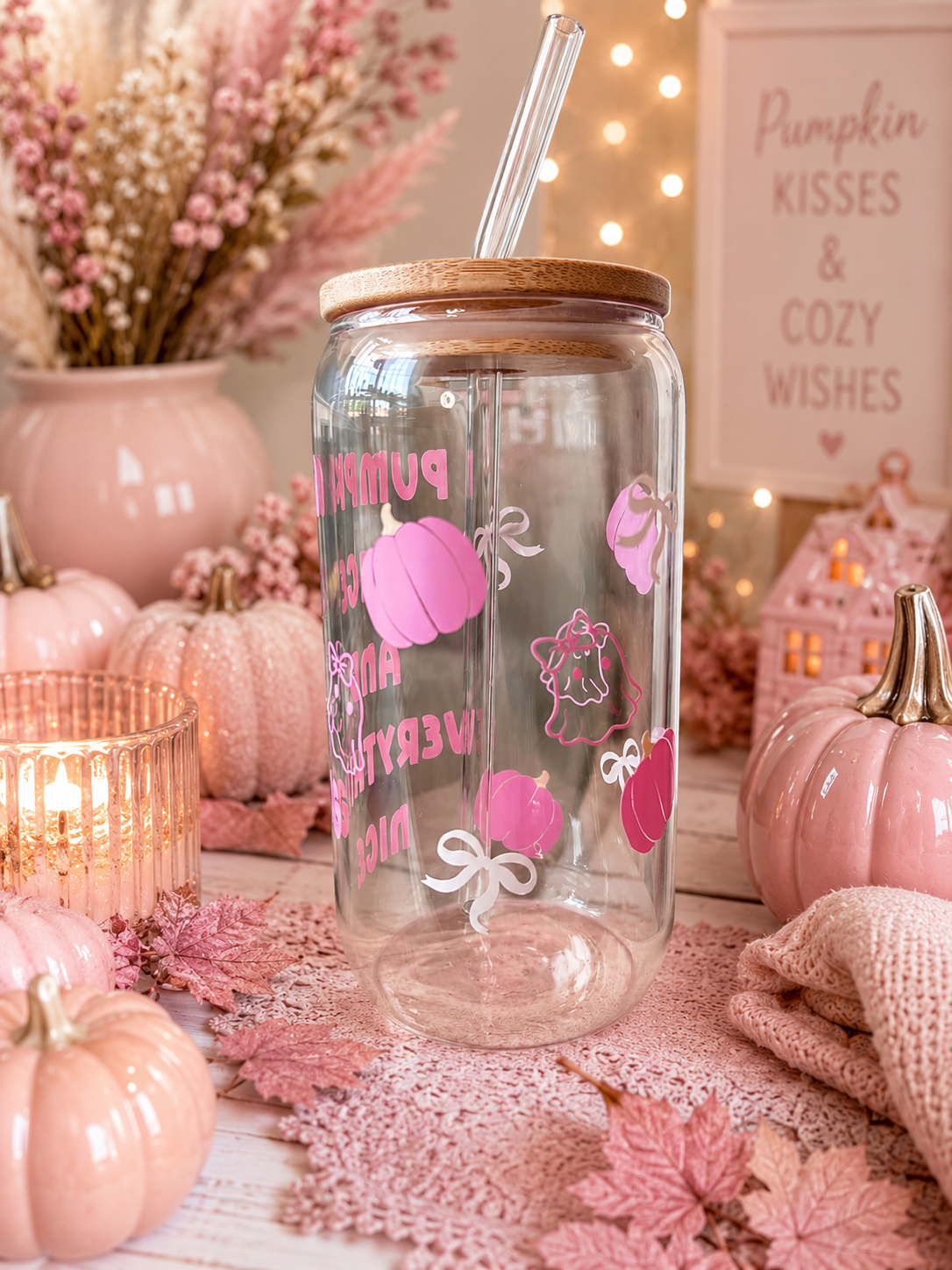 A clear glass jar tumbler with a bamboo lid and clear straw, decorated with pink pumpkins, bows, and the words 'Pumpkin Spice and Everything Nice' in pink. The tumbler is styled in a cozy autumn setting with pink pumpkins, a candle, dried flowers, and a sign that reads 'Pumpkin Kisses & Cozy Wishes'.