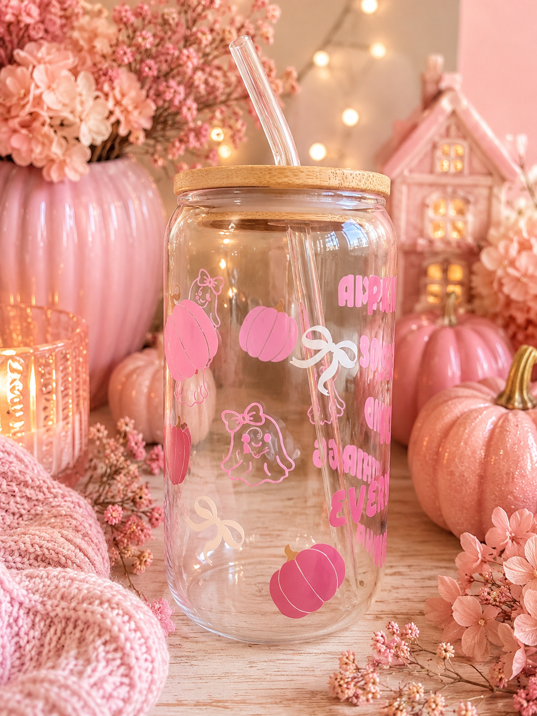 A clear glass can-style tumbler with a bamboo lid and clear straw, decorated with pink pumpkins, bows, and autumn-themed text, styled in a cozy fall setting with pink pumpkins, flowers, and string lights.