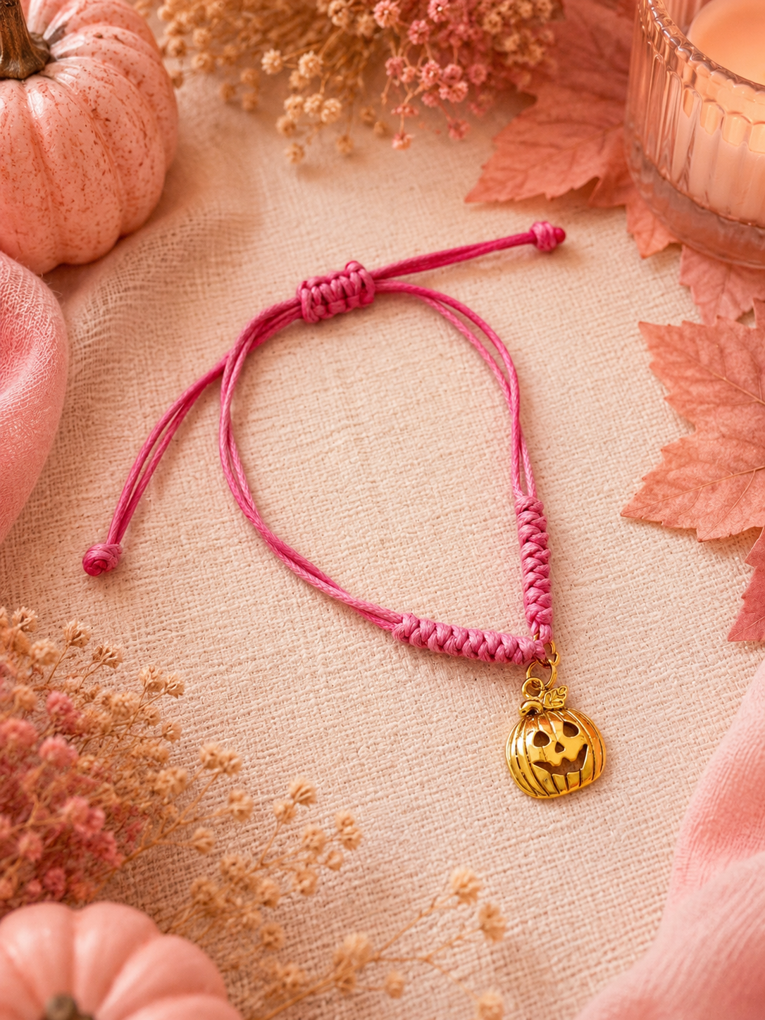 A pink cord bracelet with a small gold jack-o'-lantern pumpkin charm, styled on a beige linen fabric with autumn leaves, dried flowers, and pumpkins.