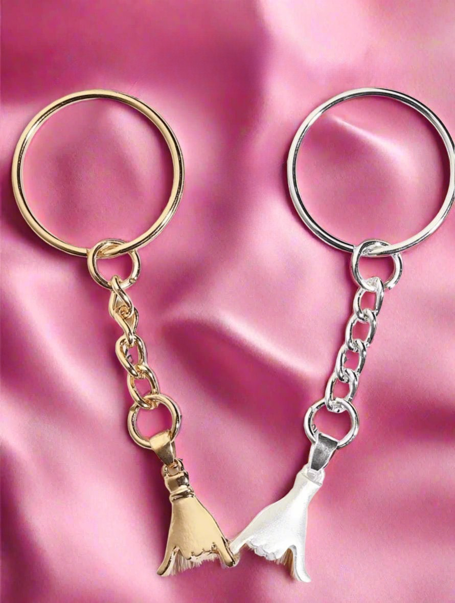 Two keychains with gold and silver pinky fingers charms on a pink fabric background