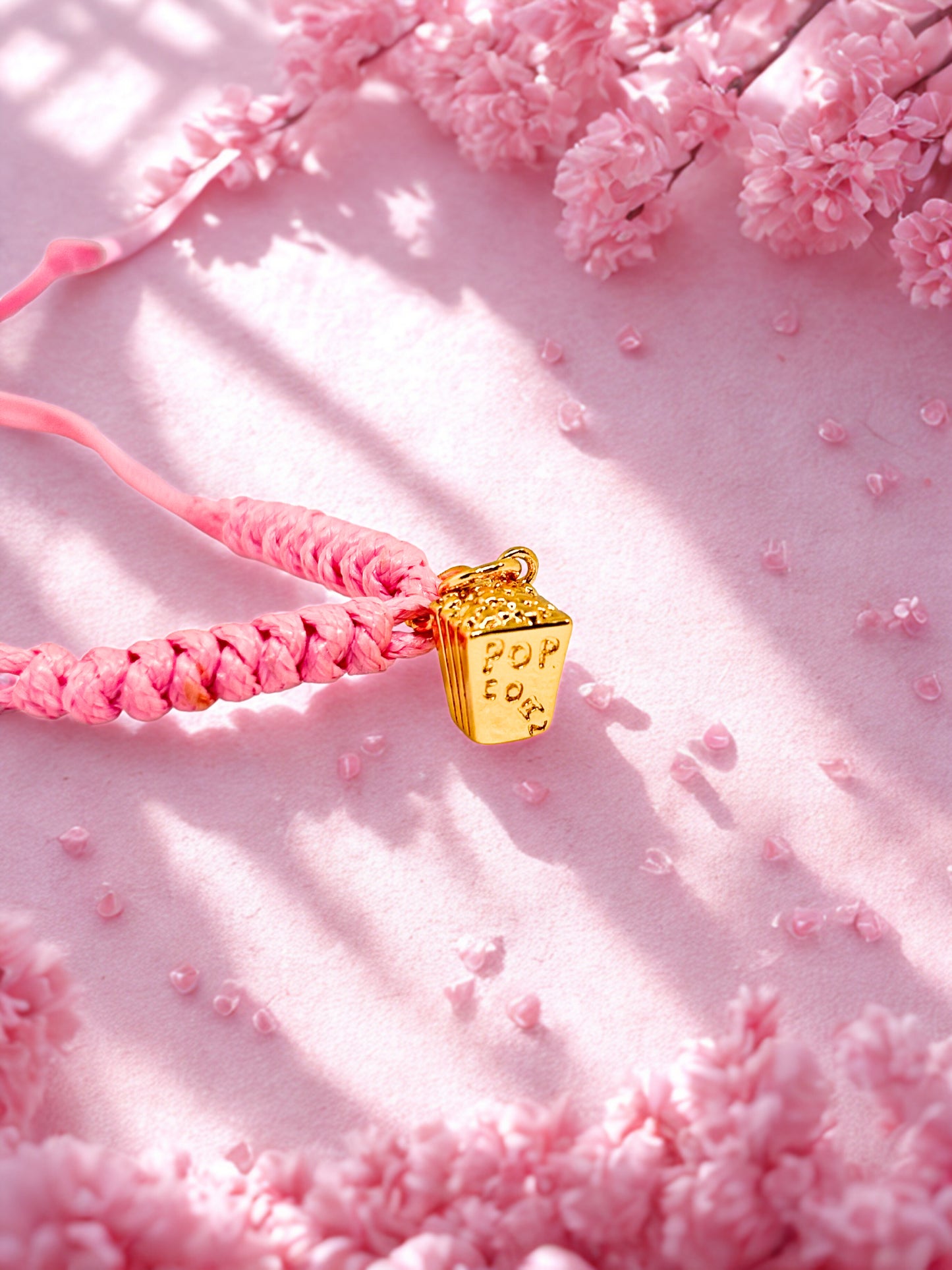 Pink braided cord bracelet with a gold popocorn charm on a pink floral background