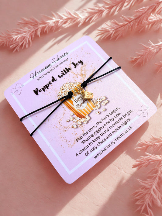 Decorative popcorn charm bracelet on a card with text, placed on a pink surface with decorative elements.