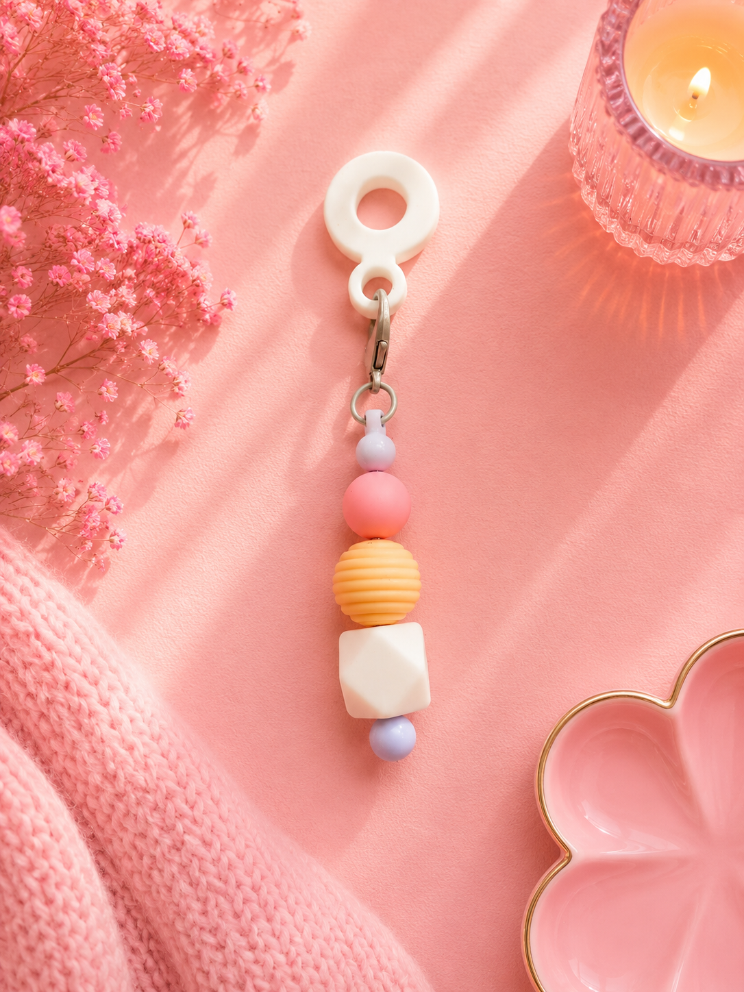 A pastel-colored beaded cup charm with a white plastic clip, displayed on a pink fabric background with decorative props including a pink candle, dried flowers, and a pink knit blanket.