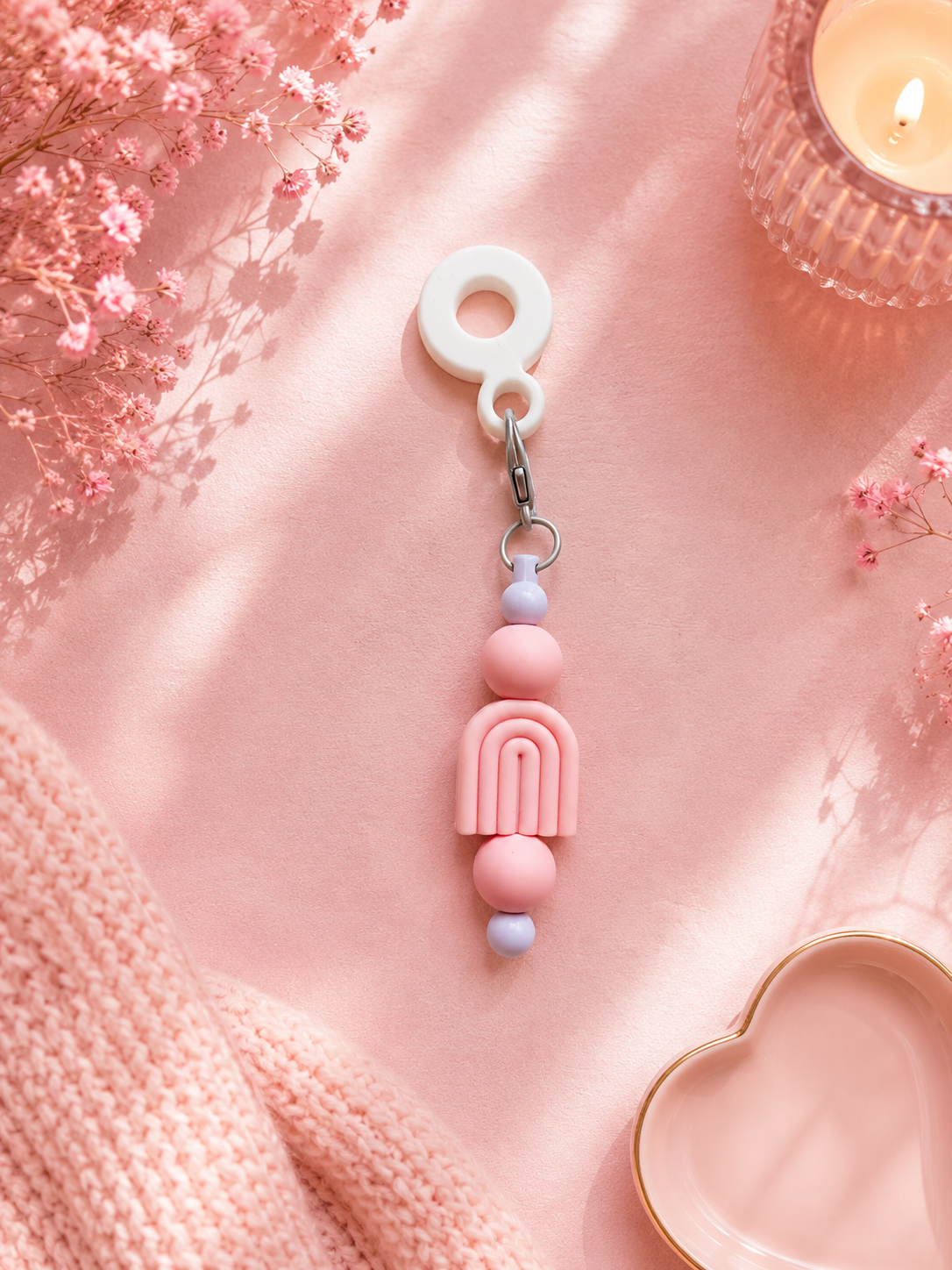 A pastel pink and white beaded cup charm with a white ring, silver metal clasp, and silicon rainbow beads detail, styled on a pink fabric background with dried flowers, a candle, and a knit textile.