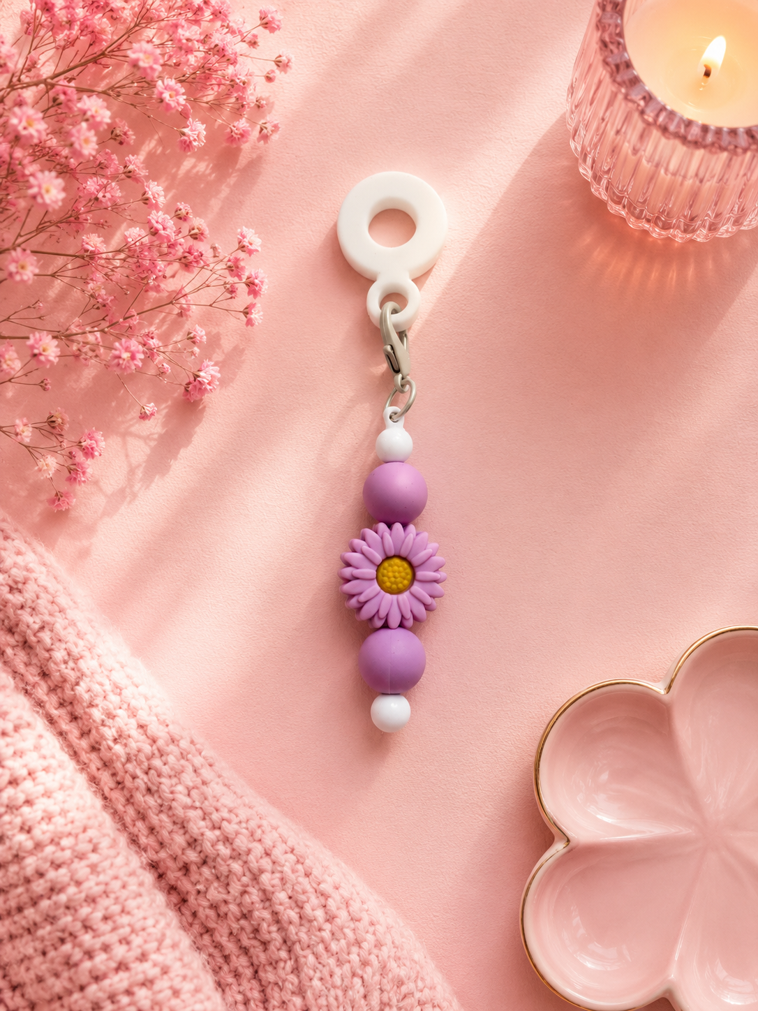 A purple and lavender beaded cup charm with a daisy flower in the center, hanging from a white ring on a pink fabric background with decorative props including pink flowers, a candle, and a pink knit blanket.