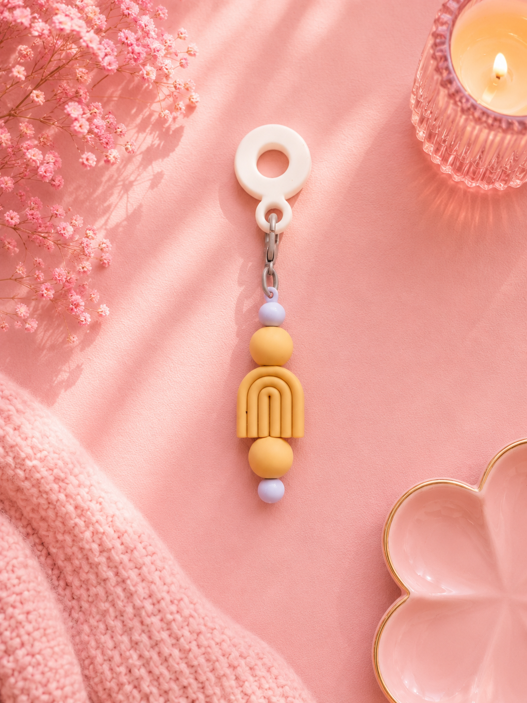 A pastel lemon beaded cup charm with a white ring, styled on a pink background with dried flowers, a candle, and a pink knit blanket.