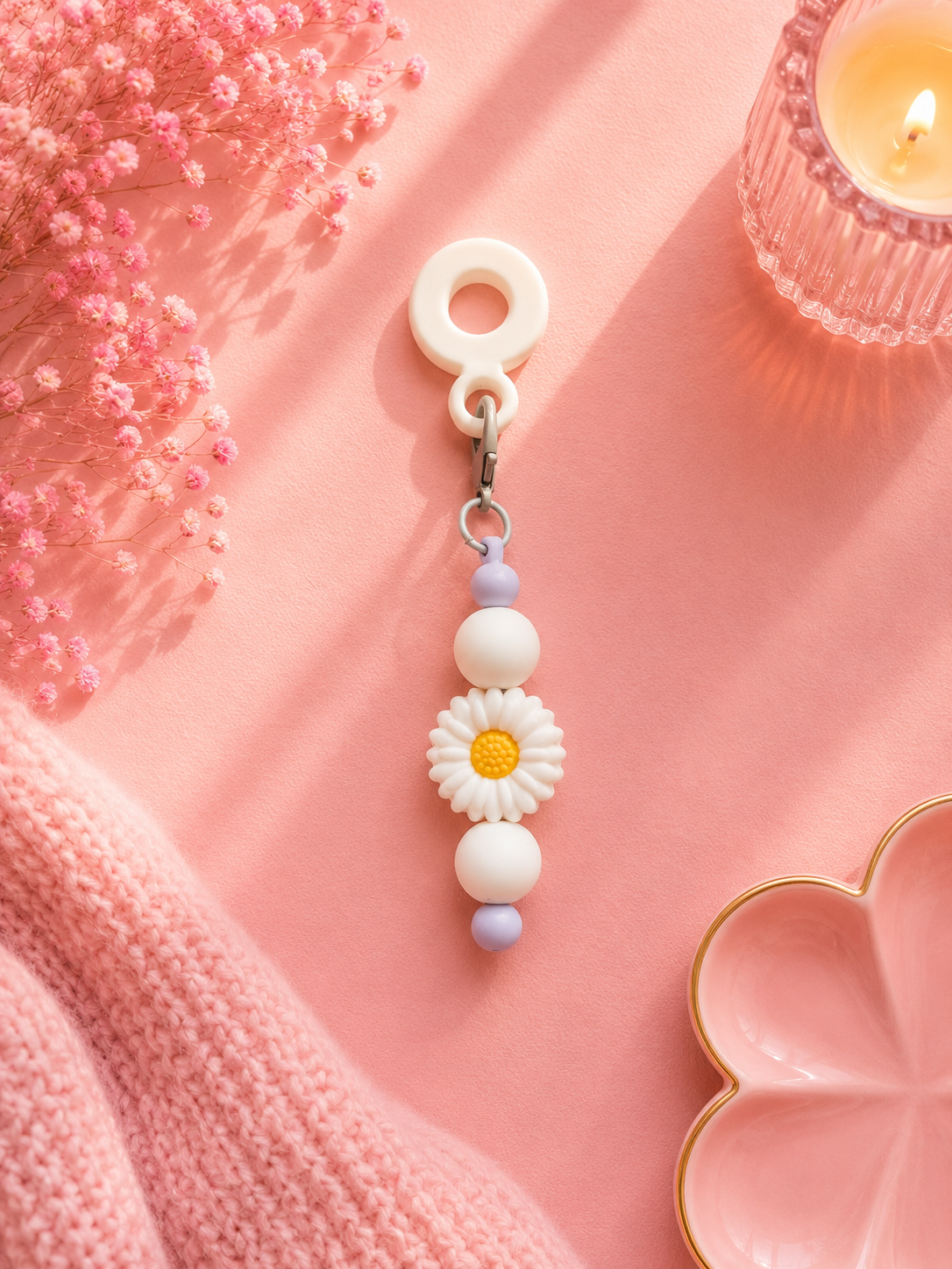A pastel white cup charm featuring a white daisy with a yellow center, styled on a pink background with a pink knit blanket, dried pink flowers, and a lit candle.