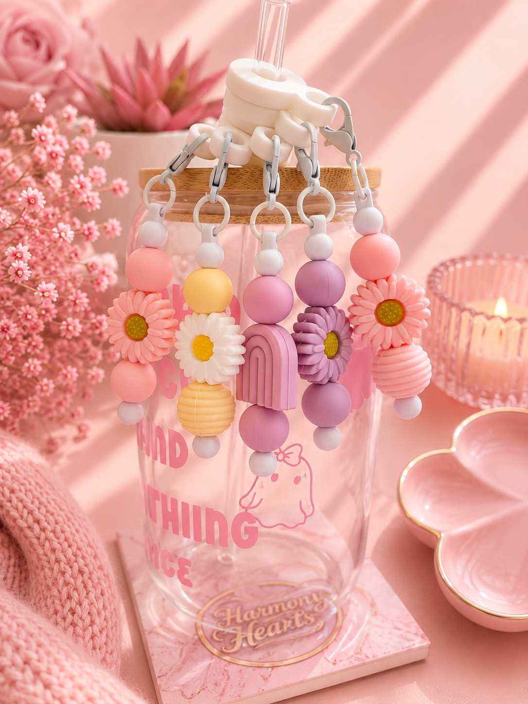 A clear glass water bottle with pink printed text and a bamboo lid, decorated with pastel flower and arch-shaped silicone charms hanging from silver clips. The bottle is styled on a pink surface with pink flowers, a candle, and a knit textile in the background.