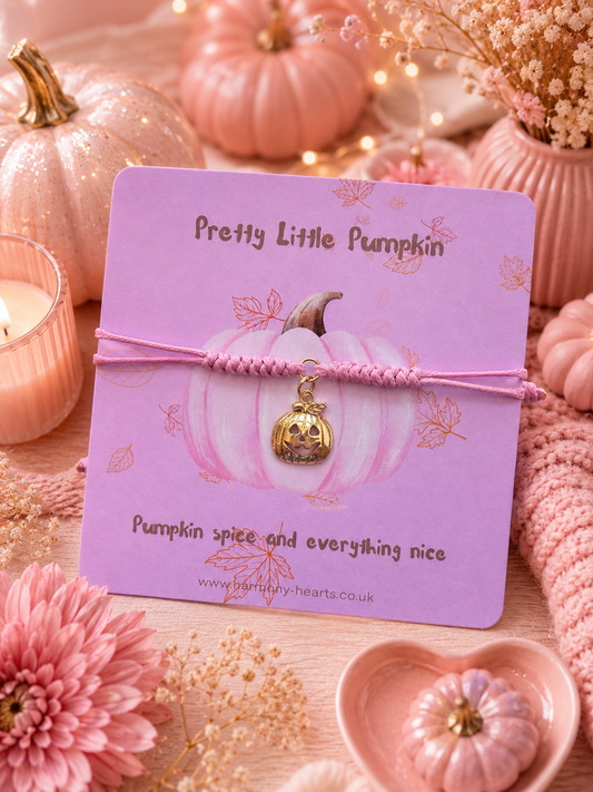 A pink cord bracelet with a carved pumpkin charm displayed on a pink card that reads 'Pretty Little Pumpkin.' The card is styled in a cozy autumn setting with pink pumpkins, flowers, a candle, and knitted textiles.