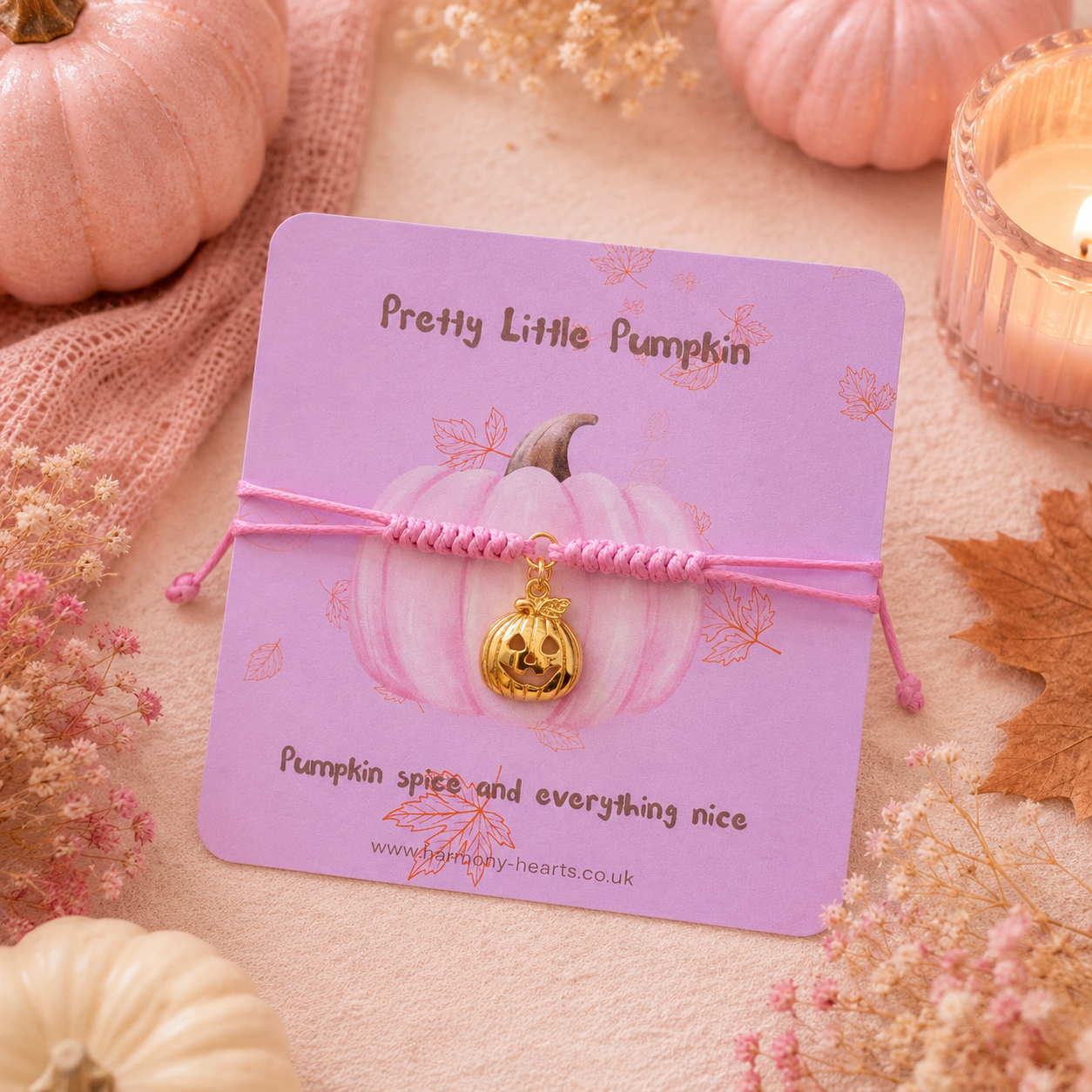 A pink cord bracelet with a carved pumpkin charm is displayed on a lavender card that reads 'Pretty Little Pumpkin.' The card is styled with pumpkins, dried flowers, and a lit candle in a warm autumn setting.