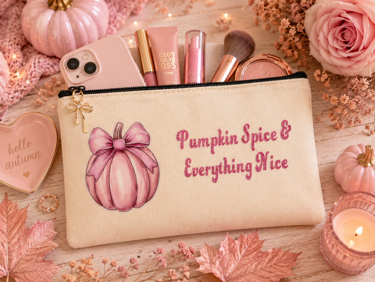 A cream-colored cosmetic pouch with a pink pumpkin illustration and the text 'Pumpkin Spice & Everything Nice' in pink script, styled on a wooden surface with pink roses, autumn leaves, a candle, and makeup items.