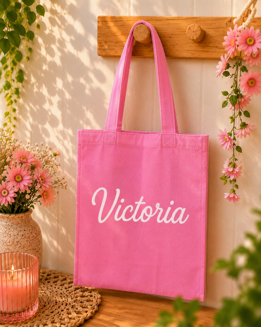 Pink tote bag with 'Victoria' printed on it, hanging on a wooden hook with flowers and a candle in the background.