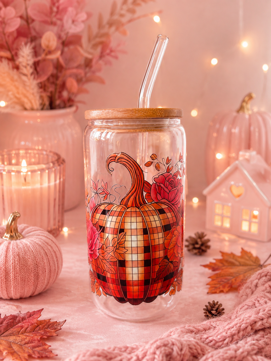 A clear glass can-shaped tumbler with a bamboo lid and glass straw, decorated with a plaid pumpkin and autumn leaves. The tumbler is styled in a cozy, warm fall setting with pink knit textures, candles, pinecones, and a small house ornament.