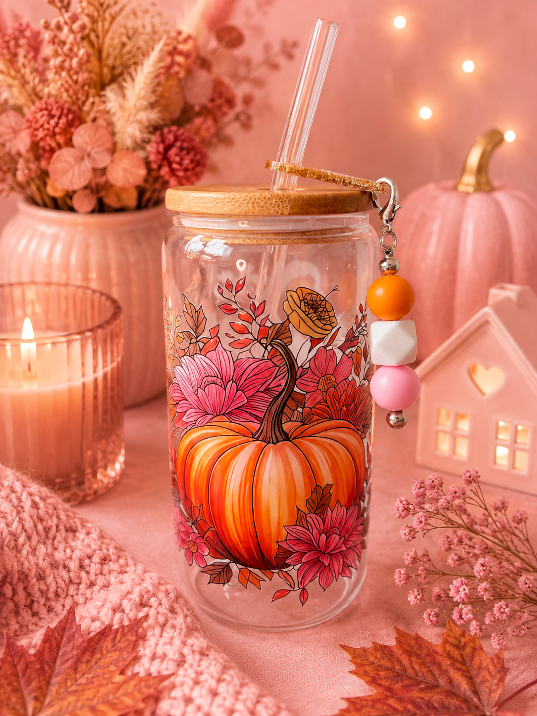 A clear glass  tumbler with a bamboo lid and straw, decorated with a large orange pumpkin and pink floral design. It features a decorative beaded charm.
