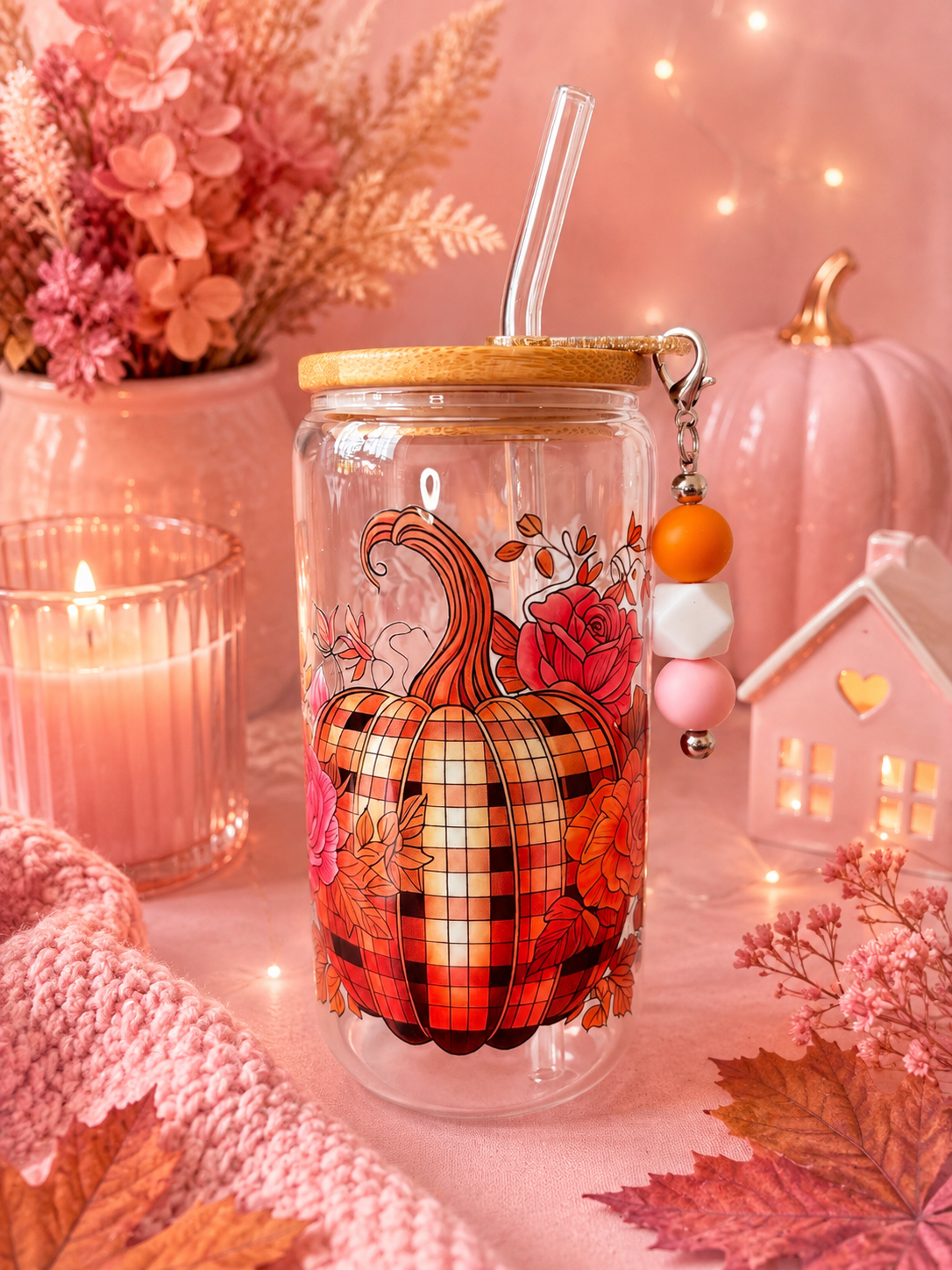 A clear glass tumbler with a bamboo lid and clear straw, decorated with a large orange and red plaid pumpkin and floral motifs, styled in a cozy autumn setting with a candle, pink pumpkins, and fall leaves.