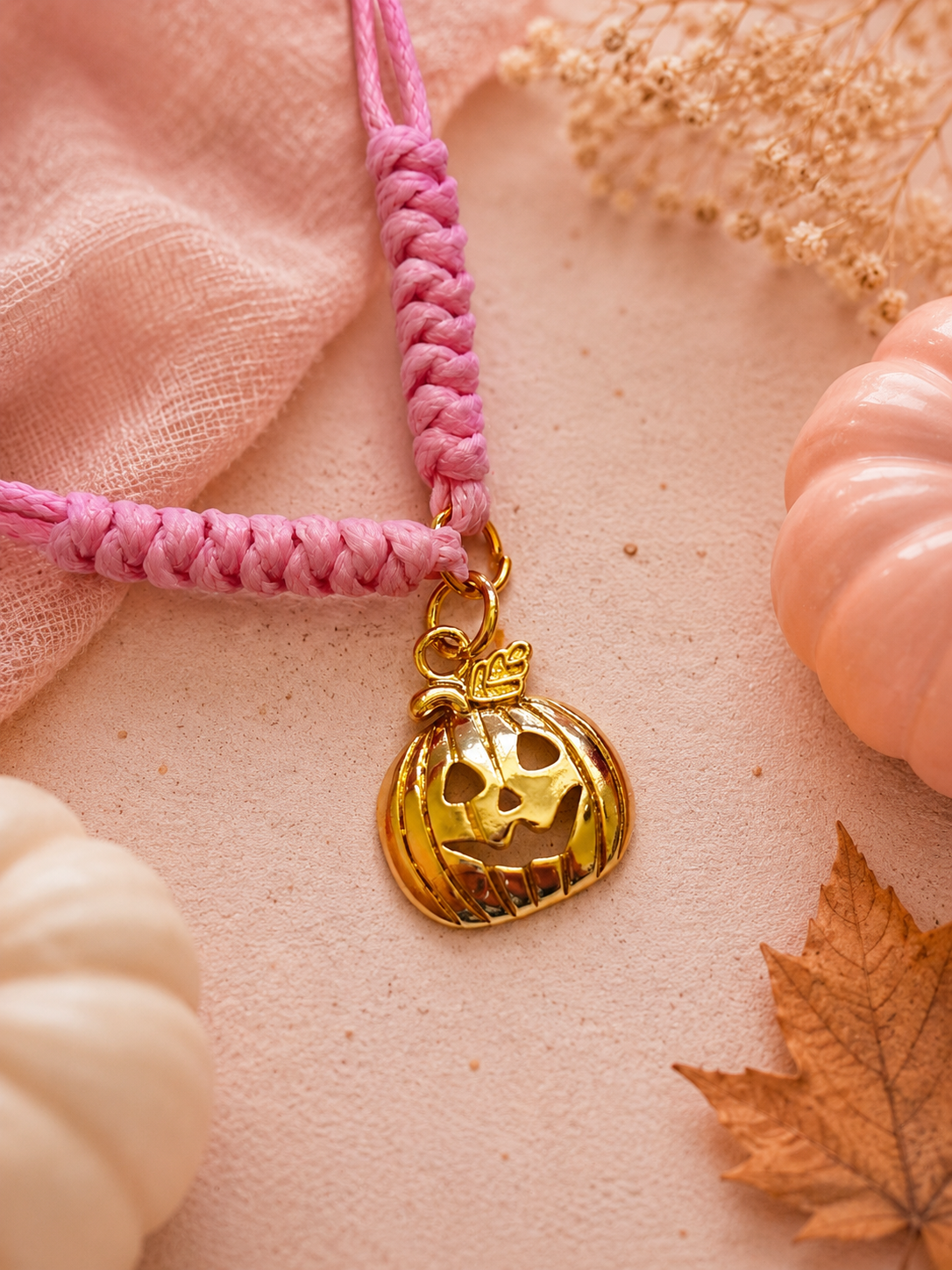 A pink braided cord bracelet with a gold jack-o'-lantern pumpkin charm, styled on a soft pink fabric background with dried flowers, autumn leaves, and small decorative pumpkins.