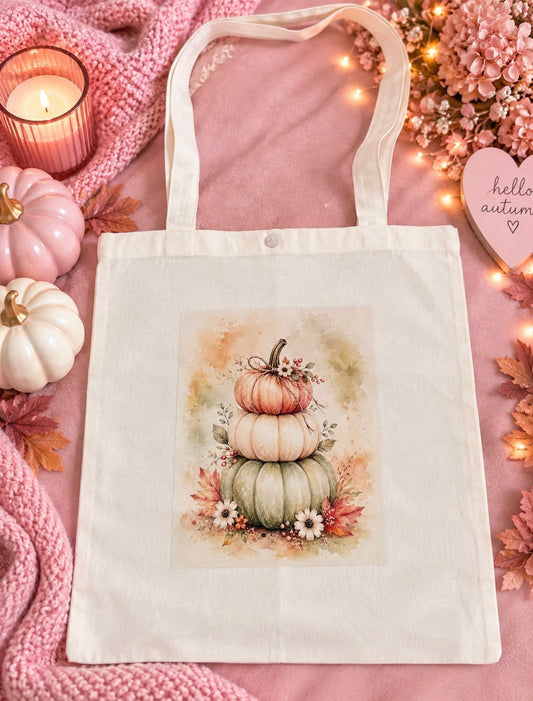 A cream canvas tote bag with a printed watercolor illustration of stacked autumn pumpkins and small white flowers, styled on a pink blanket with candles, pumpkins, leaves, and a heart-shaped sign.