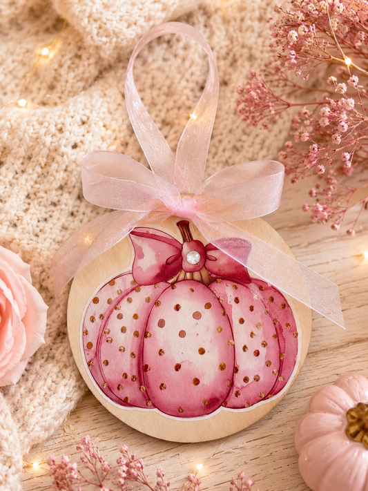A round wooden autumn ornament shaped like a pink watercolor pumpkin with gold speckles, tied with a sheer blush pink ribbon. The ornament is styled on a light wood surface with a cozy beige knit blanket, pink roses, dried baby's breath, and warm fairy lights.