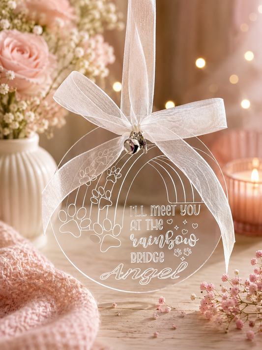 A clear acrylic hanging ornament shaped like a rainbow arch with floral line art and the phrase 'I'll meet you at the rainbow bridge Angel' printed on it. The ornament is tied with a sheer white ribbon and a small silver bell, shown in a soft pink styled setting with flowers, a candle, and a knitted textile.