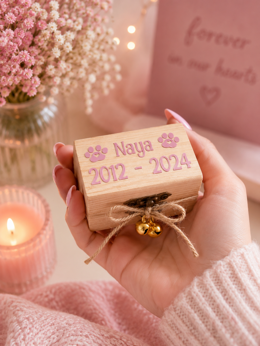 A hand holds a small wooden keepsake box with the name 'Naya', dates '2012 - 2024', and paw prints, tied with twine and small gold bells. The background is softly blurred with pink flowers, a lit candle, and a pink memorial sign reading 'forever in our hearts'.