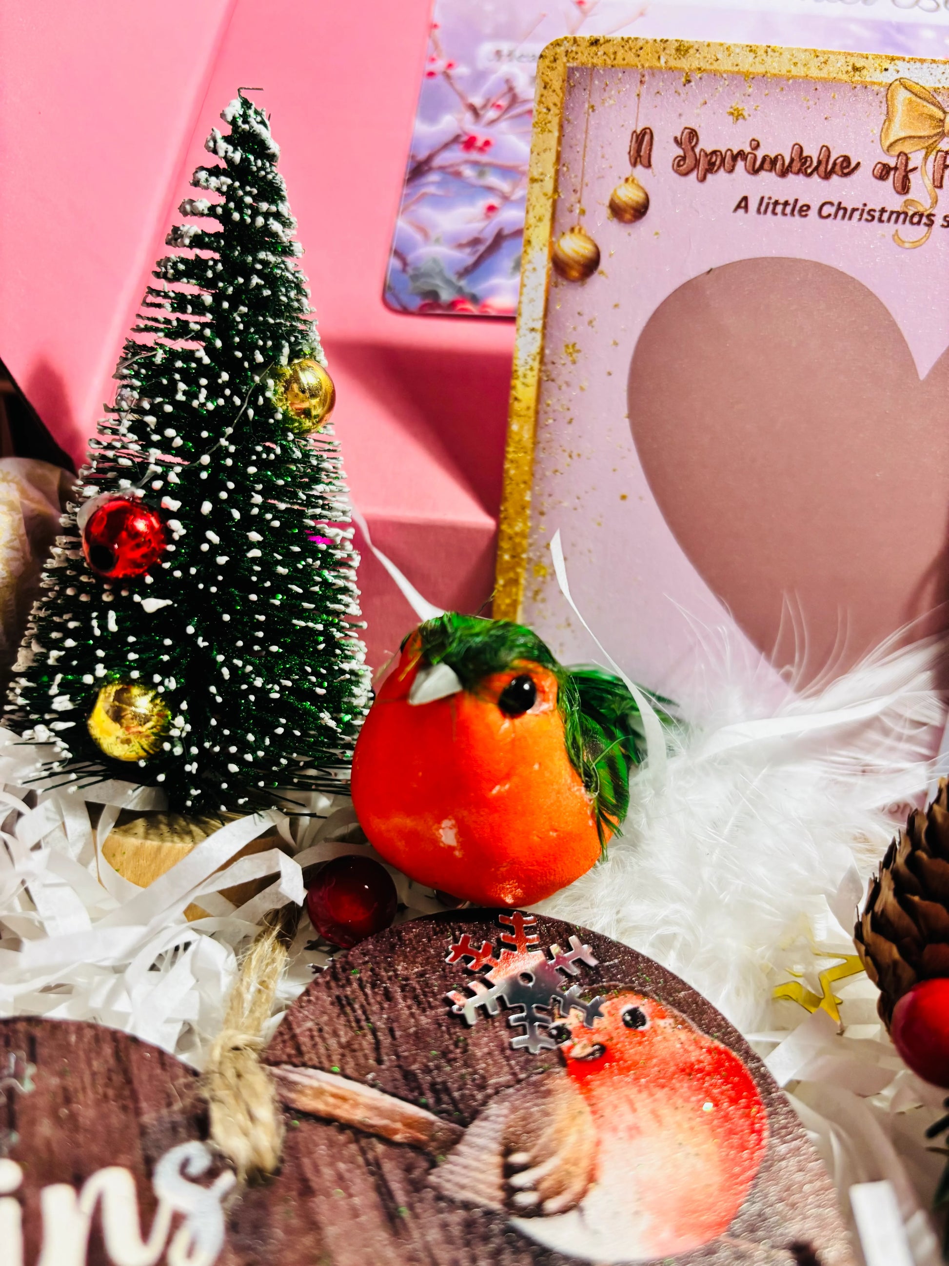 Decorative Christmas scene with a small tree, robin, and festive card on a pink background.