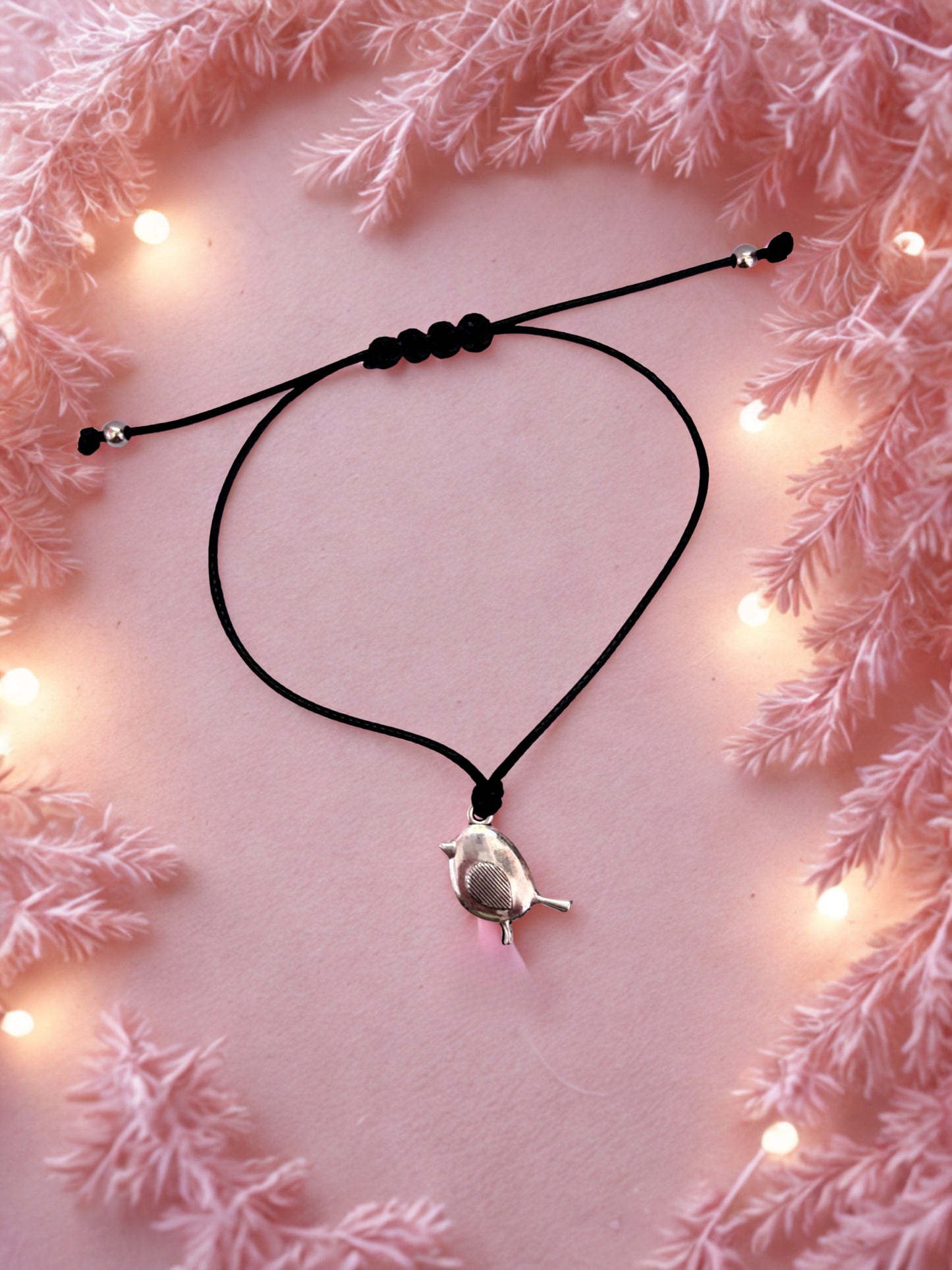 Black cord bracelet with a robin  charm on a pink surface with decorative lights and branches.