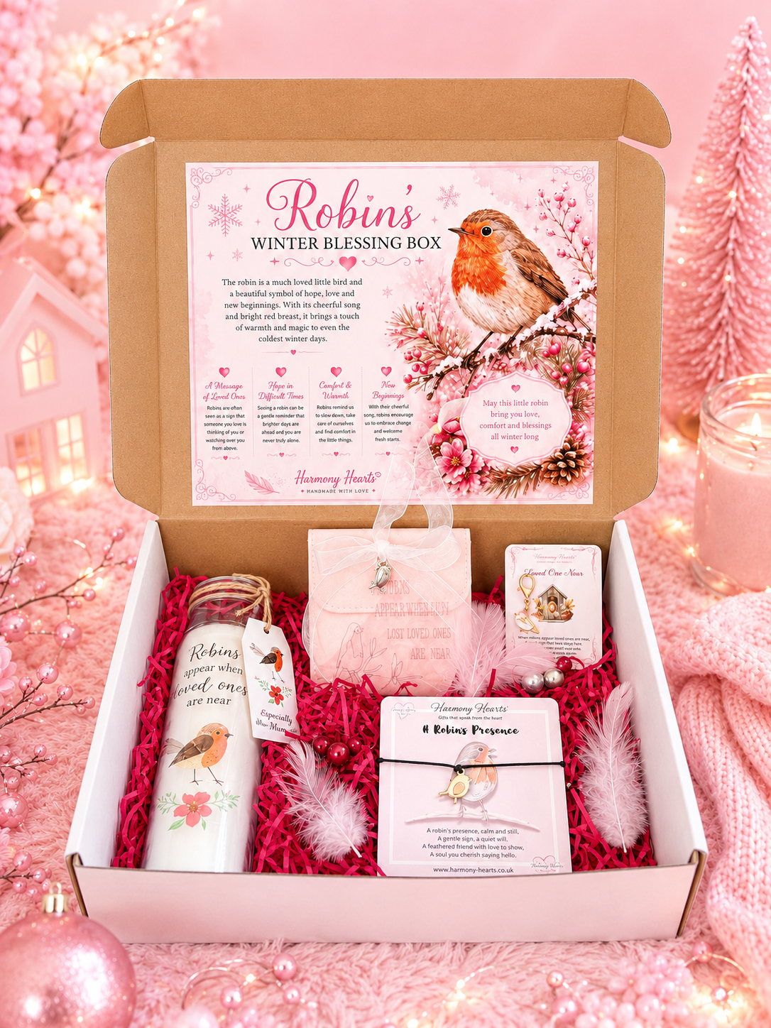 An open cardboard gift box labeled 'Robins' Winter Blessing Box' filled with pink shredded paper and holiday-themed items, including a white candle with robin illustrations, a small pink pouch, cards, and feathers, set against a soft pink festive background.