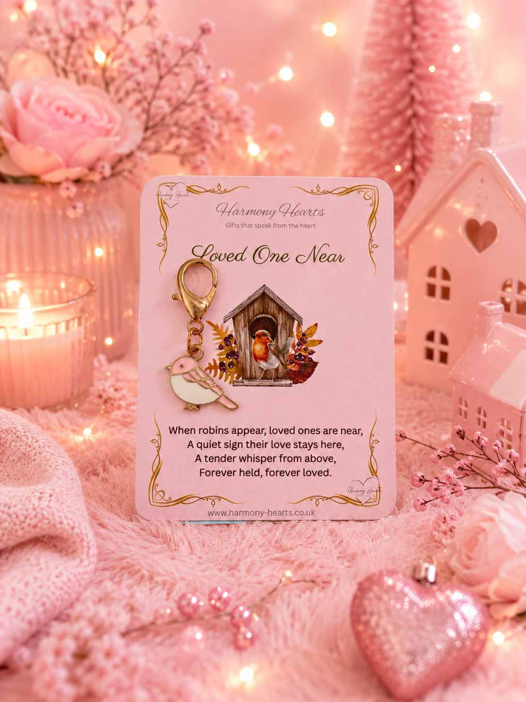 A pink memorial card with a robin keychain charm, displayed in a cozy pink-themed setting with candles, fairy lights, and a small house.