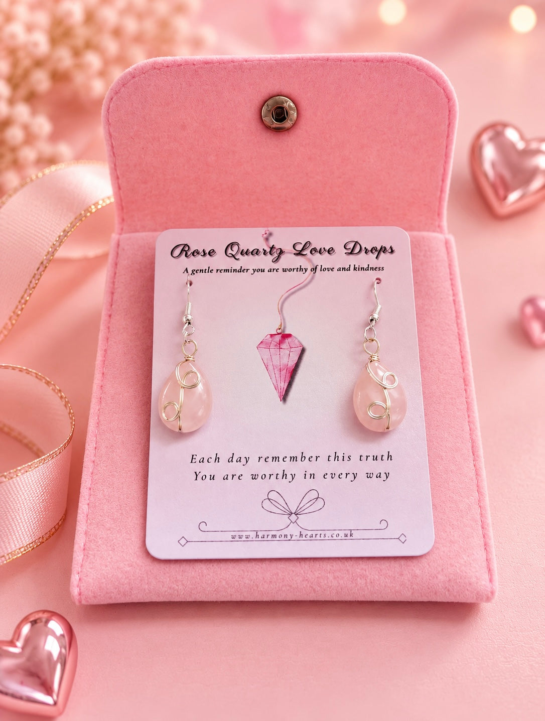 A pair of rose quartz love drop earrings displayed on a white branded card inside an open pink velvet jewelry box, with pink heart decorations and ribbon around it.