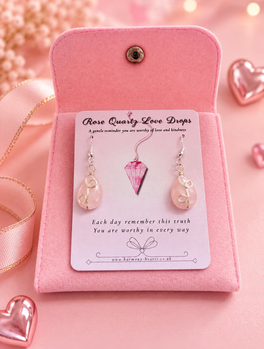 A pair of rose quartz love drop earrings displayed on a white branded card inside an open pink velvet jewelry box, with pink heart decorations and ribbon around it.