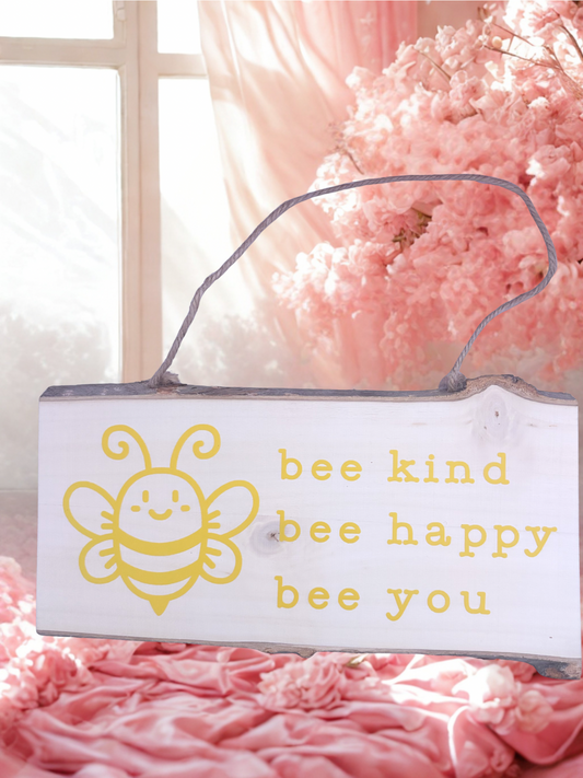 Decorative sign with a bee illustration and text 'bee kind bee happy bee you' on a pink floral background.