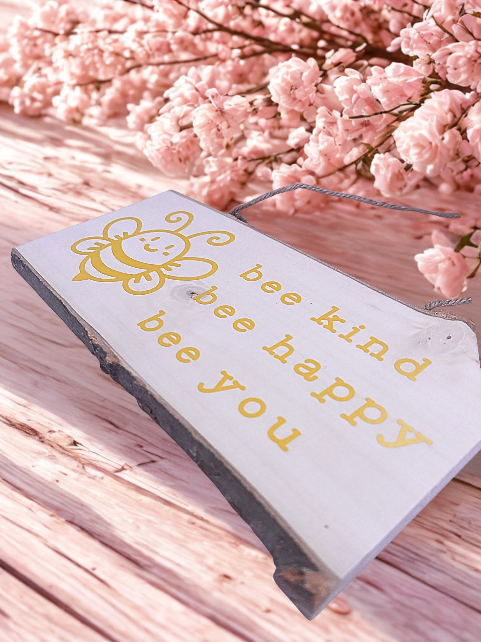 Decorative sign with 'bee kind bee happy bee you' text on a wooden surface with pink flowers in the background.