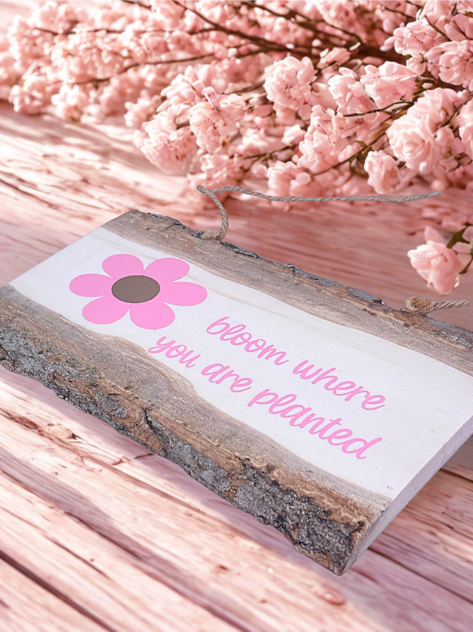 Decorative sign with pink flower and text 'bloom where you are planted' on a wooden background with pink flowers.