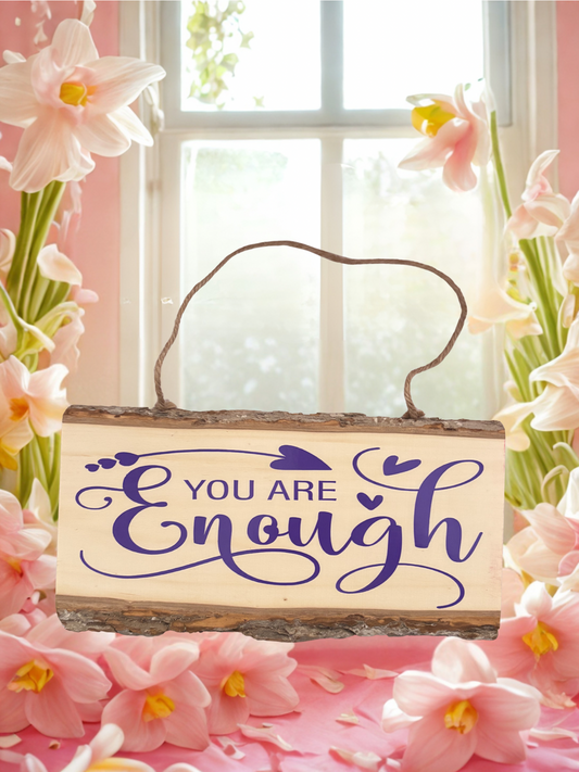 Wooden sign with 'You Are Enough' text surrounded by pink flowers in front of a window.