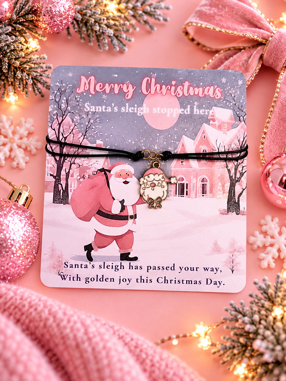A Christmas-themed black cord bracelet with a Santa charm, displayed on a printed holiday card. The card is styled on a pink background with festive ornaments, pine branches, and lights.