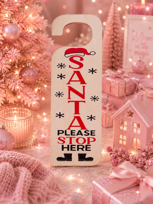 A Christmas-themed wooden door hanger sign with the text 'SANTA PLEASE STOP HERE' in red and black, surrounded by pink holiday decorations, gift boxes, a lit candle, and a small pink house ornament.