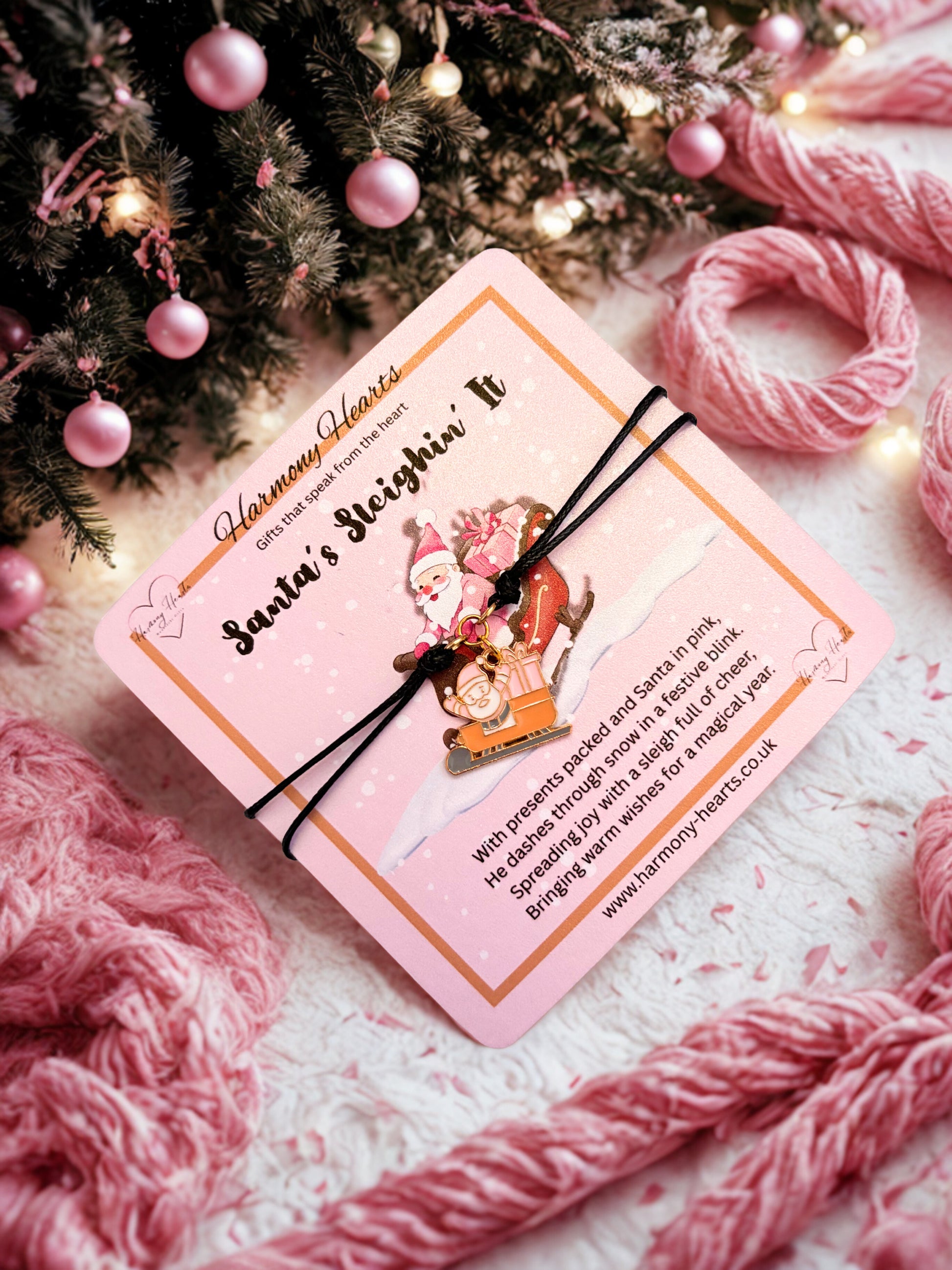 Black cord charm bracelet on a pink card with Santa Claus illustration on a festive background with pink yarn and Christmas tree.