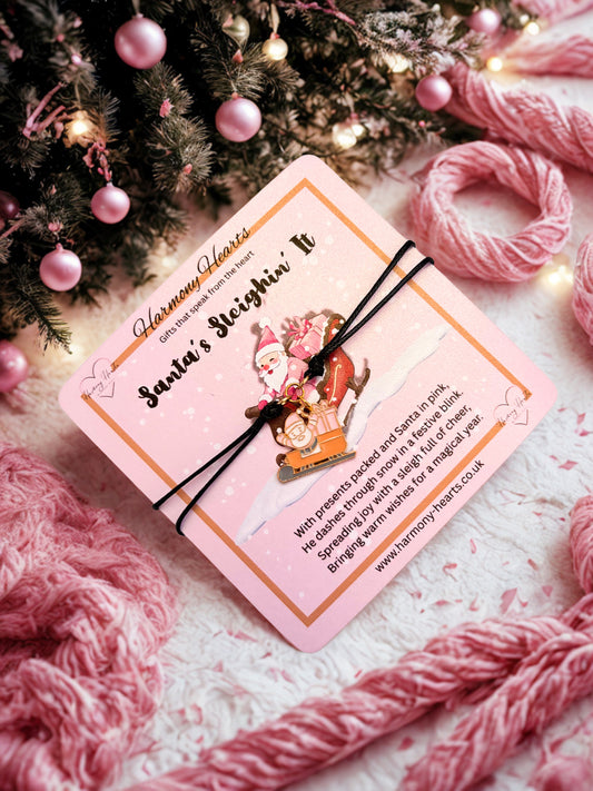 Black cord charm bracelet on a pink card with Santa Claus illustration on a festive background with pink yarn and Christmas tree.