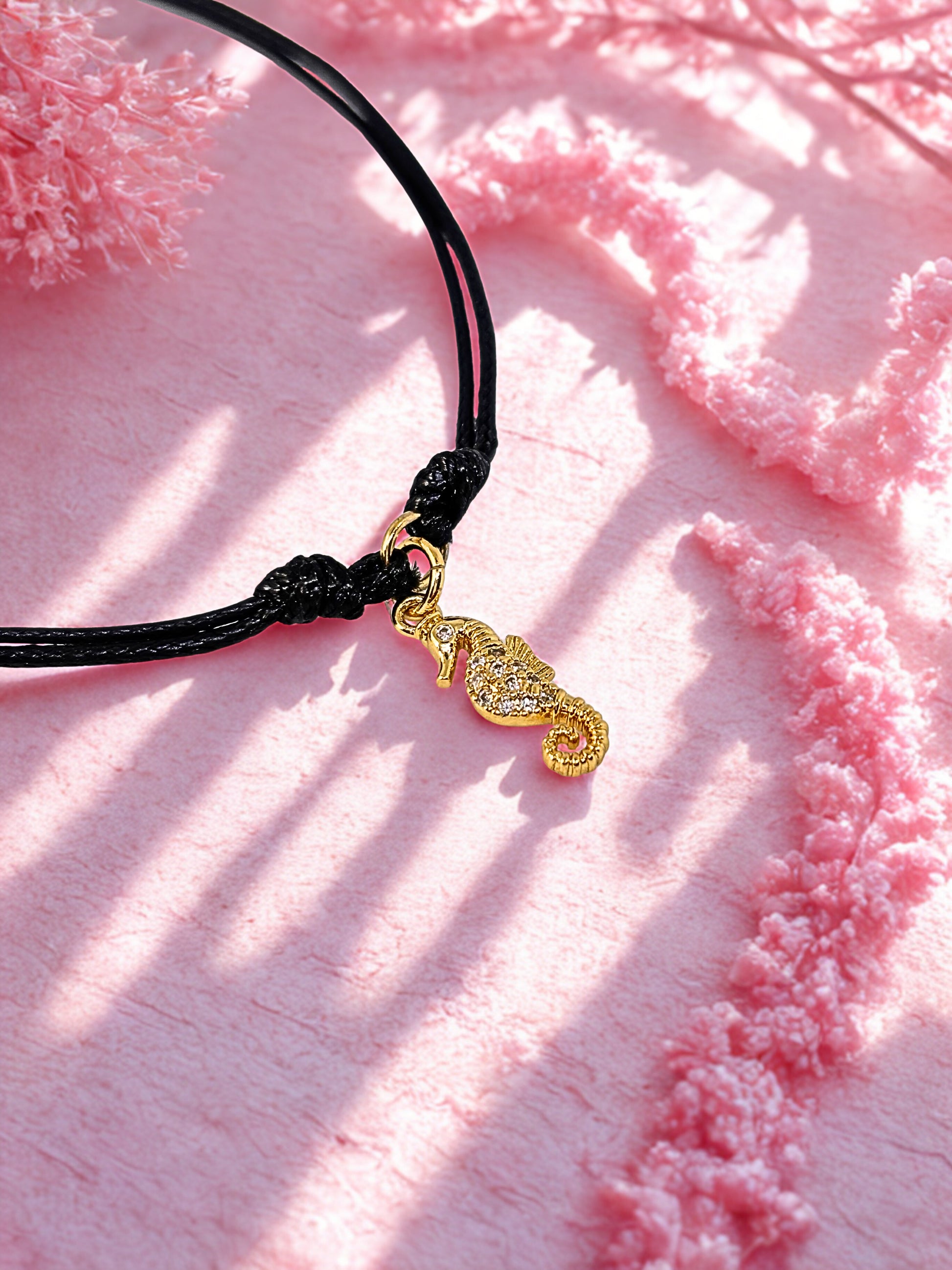 Black cord bracelet with a gold seahorse charm on a pink textured background